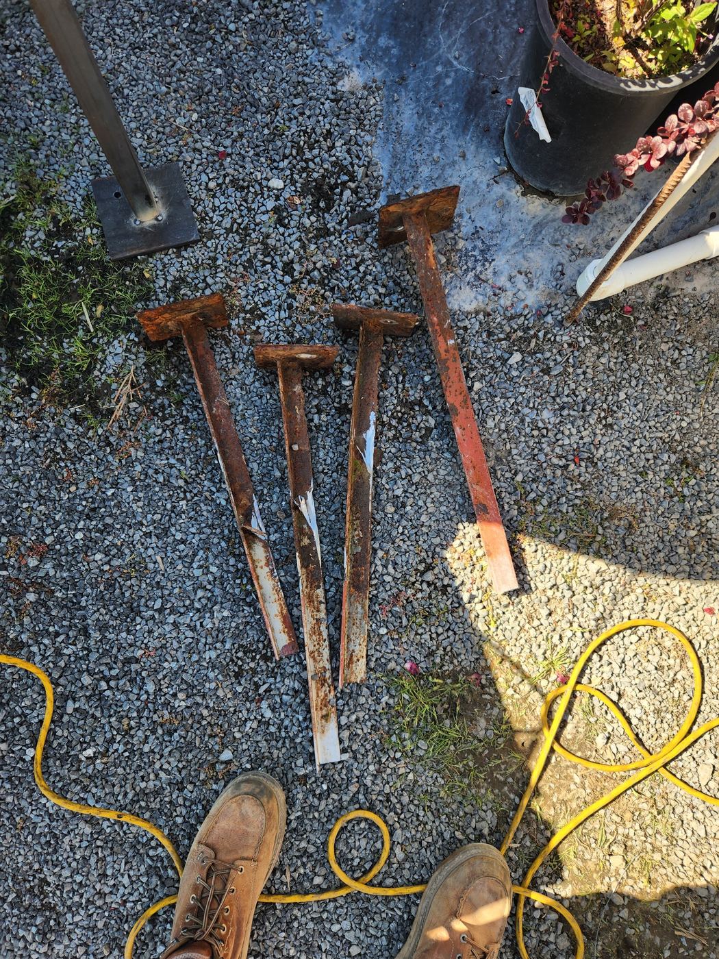 Four removed rusted angle-iron post stubs laid out on gravel after the repair